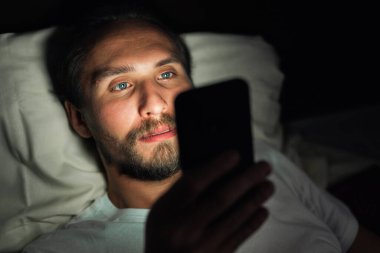 Sakallı, genç, yakışıklı ve yorgun bir adam uyuyamıyor ve geceleri telefonundan bir şey izliyor..