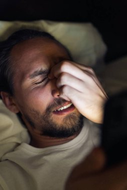 Sakallı beyaz erkek gece yatağında telefonundan bir şey izliyor. Yorgun, uyuyamıyor ve belli ki başı ağrıyor..