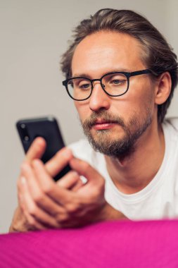 Yatakta yatarken mobil cihazıyla etkileşime geçen oldukça ciddi bir genç adamın fotoğrafı.