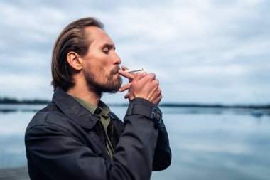 Sigara içen genç sakallı bir adamın fotoğrafı. Arka planda göl ve bulutlar.