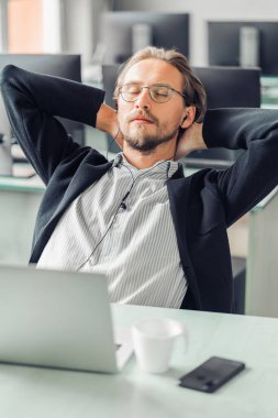 Genç adam iş yerinde müzik dinlerken dinlenmeye ve uyumaya çalışıyor. Kahve fincanı, bilgisayar ve ön planda görünür bir cep telefonu..
