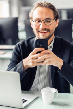 Young handsome man in glasses is interacting with his mobile phone while being at work. Happiness and content at work and being always reachable concepts. clipart