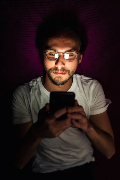一个留着胡子的年轻人晚上躺在床上 一边看手机上的东西 男人的眼镜上可以看到手机的倒影 — 图库照片