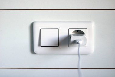 Avrupa prizine takılmış elektronik bir cihazın şarjı. Avrupa tıkacı ve beyaz ışık düğmesi.