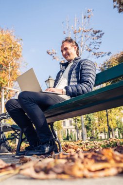 Genç bir adam güzel bir sonbahar gününde bankta oturuyor. Taşınabilir bilgisayarını kullanıyor..