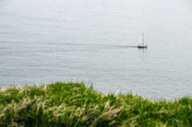 Sakin deniz yüzeyinde huzurlu bir yelkenli manzarası ve bulutlu bir günde yeşil tepe.