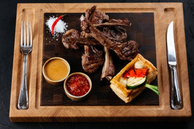 Izgara kuzu pirzola, sebzeli ve soslu siyah arka plan.