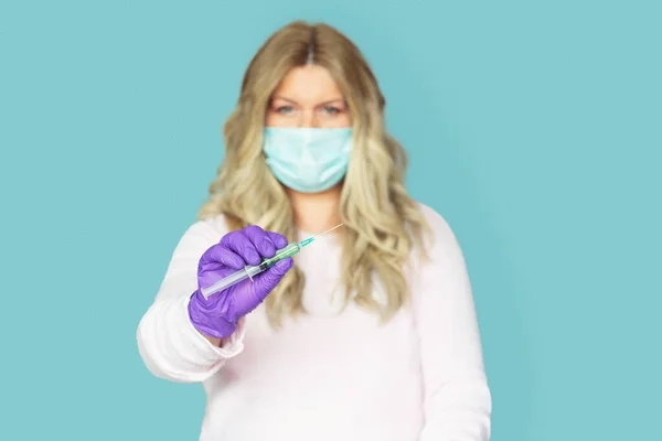 BLONDE WOMAN WITH GLOVES AND MASK HOLDING A SYRINGE WITH A VACCINE IN HER HAND