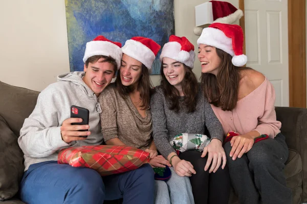 Four college-aged siblings wearing Santa hats look at selfies on