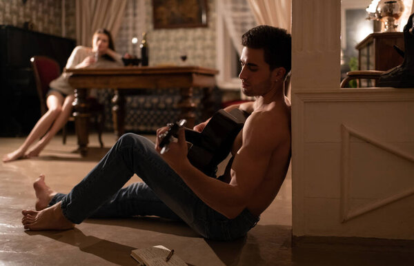 Side view of barefoot male musician sitting on floor in old apartment and playing acoustic guitar while creating song for blurred girlfriend during romantic date