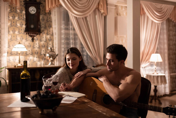 Serious young man with guitar making notes in planner while sitting near cheerful girlfriend and creating song during romantic date in aged apartment
