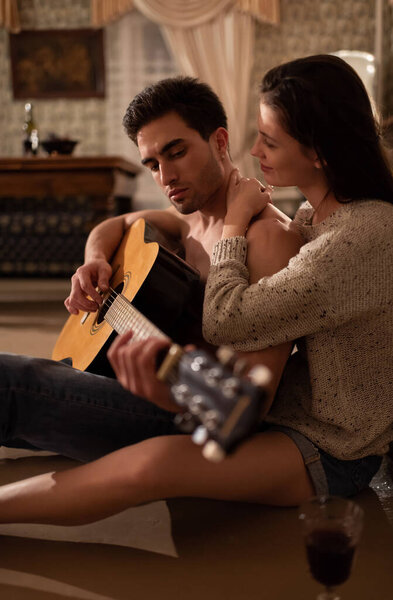 Side view of glad young female smiling and embracing boyfriend while sitting on floor near glass of wine and listening to romantic song in retro apartment
