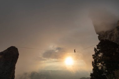 Bir kişi bulutlu bir gün batımında büyük kayaların arasında yüksek bir çizgide yürüyor.