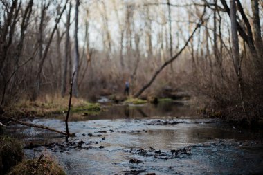 Önünde karanlık ve kasvetli bir dere olan bir adam arka planda duruyor.