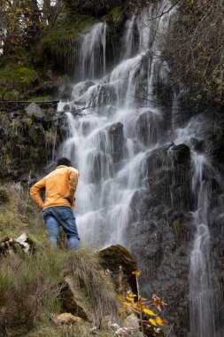 Bir adam kayanın üzerinde oturuyor ve arka planda küçük bir şelale ve sırt çantasıyla uzaklara bakıyor.
