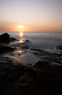 Sahil şeridinin uzun pozlama manzarası: sudaki günbatımının renkleri kayalar ve gökyüzü