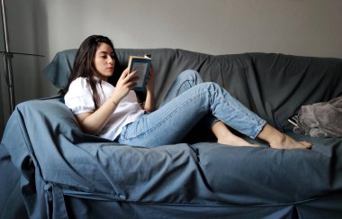 Evdeki kanepede kitap okuyan bir kadın.