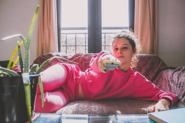 Genç bir kadın televizyon kumandasıyla kanepede uzanıyor.
