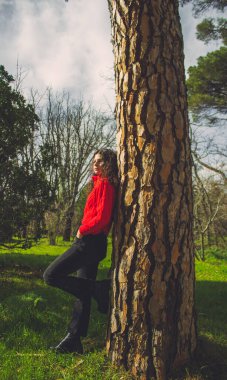 Ormandaki bir ağaca yaslanmış kırmızı kazaklı genç bir kadın.