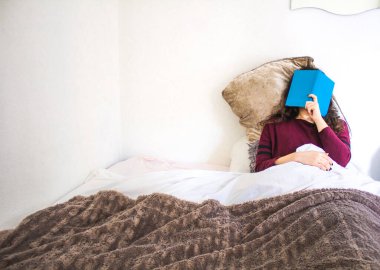 Genç bir kadın, mavi bir kitapla yatakta uzanıyor.