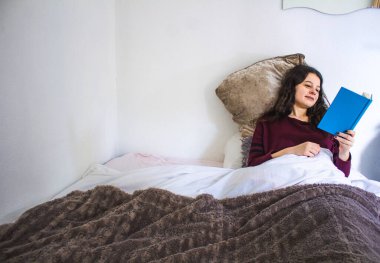 Genç bir kadın, mavi bir kitapla yatakta uzanıyor.