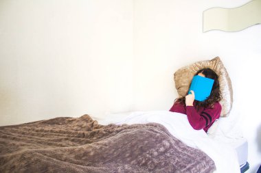 Genç kadın yatakta uzanıp kitap okuyor.