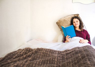 Genç kadın yatakta uzanıp kitap okuyor.