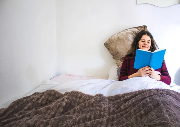 一个年轻女人躺在床上看书 手里拿着一本蓝皮书 — 图库照片