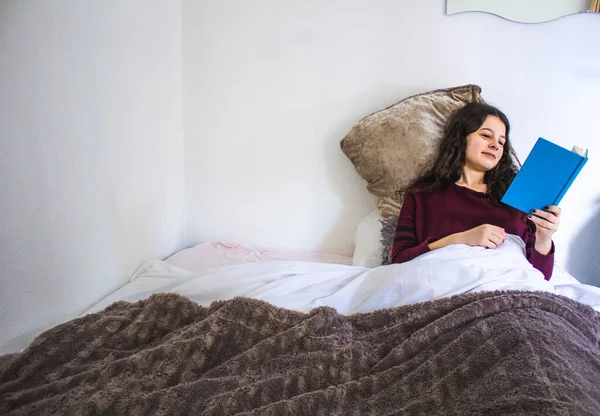 Genç bir kadın, mavi bir kitapla yatakta uzanıyor.