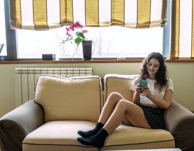 Genç bir kadın cep telefonuyla kanepede uzanıyor.