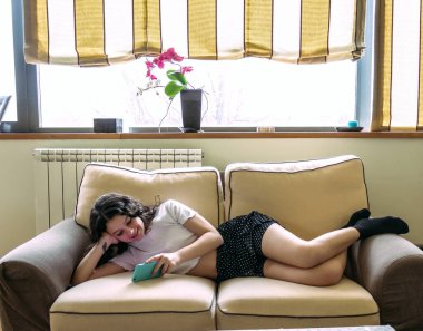 Genç bir kadın cep telefonuyla kanepede uzanıyor.