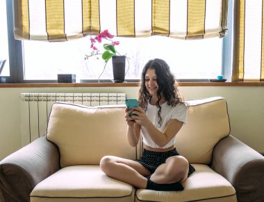 Genç bir kadın cep telefonuyla kanepede uzanıyor.