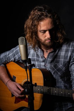 Çekici bir adam mikrofonun yanında oturup akustik gitar çalıyor.