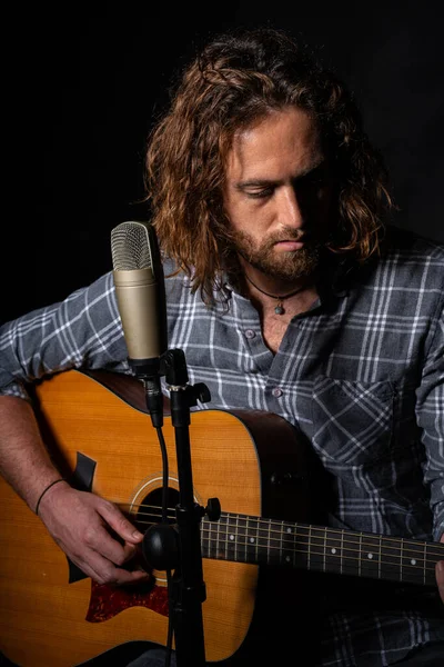 Çekici bir adam mikrofonun yanında oturup akustik gitar çalıyor.