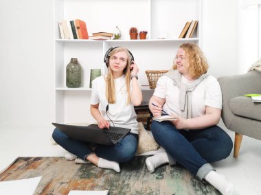 İki kadın yerde dizüstü bilgisayar ve kulaklıklarla ders çalışıyor.