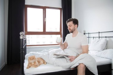 Genç bir adam gazete okur ve yatakta kahve içer, gerçek hayatta, kedi