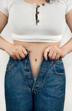 Beyaz bluzlu genç kadın kot pantolon iliklemeye çalışıyor.