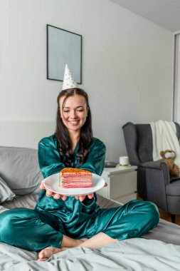 Genç bir kadın pijamalarıyla yatağa oturur ve bir tabakta kek uzatır. Sağlık konsepti. Tatilde evde kal.