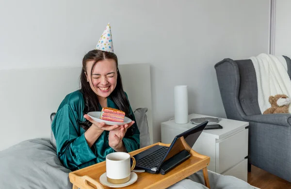 Young woman communicates with friends the Internet, using the computer, sitting the bed the bedroom. Celebrating birthday online. People wellness concept. Comfort zone in the house.