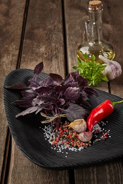 Fresh purple basil with garlic, red hot pepper, coarse sea salt and peppercorns on black plate and decanter of olive oil on wooden table. Set of ingredients for cooking flavored sauce