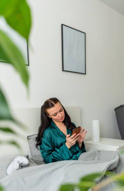 Yatakta uzanmış ipek pijama giyen genç bir kadın telefonunu tutuyor. Kendi evinizin rahatı için çalışın. Refah kavramı.