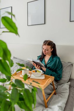 İpek pijamalı genç bir kadın yatakta yatarken sağlıklı kahvaltı yapar ve kitap okur..