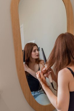 Genç kızıl saçlı kadın aynanın karşısında oturuyor ve saçını ve makyajını yapıyor. Evde bakım konsepti. Çevre dostu kozmetik ürünleri kullanımı.