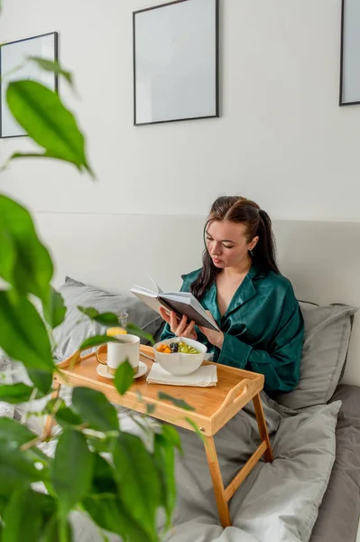 若いです女性でシルクパジャマ持っています健康的な朝食と読書本ながらベッドルームでベッドに横たわって — ストック写真