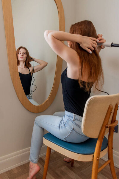 Young red-haired woman sits on chair in front of mirror doing her hair and makeup. Home care concept. Use of eco friendly cosmetics.