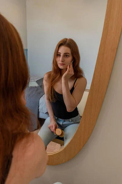 若い赤い髪の女性が鏡の前の椅子に座って髪とメイクをしています ホームケアの概念 環境に優しい化粧品の使用 — ストック写真