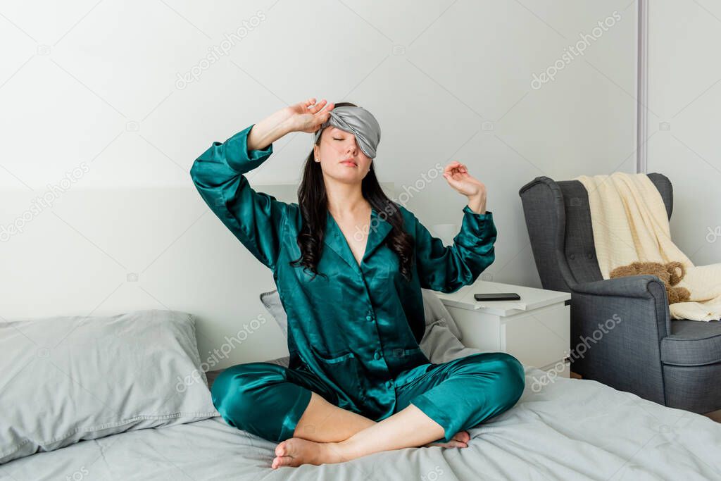 Young woman in silk pajama yawns while sitting in comfortable soft bed.