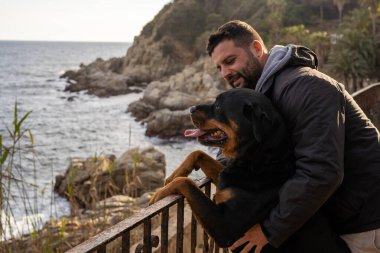 Rottweiler köpeğiyle deniz manzarasının tadını çıkaran bir adam.