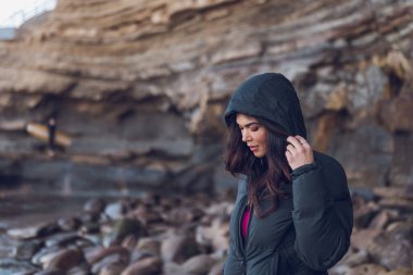 Plaj kayalıklarında sörfçülerle birlikte duran siyah kapüşonlu bir kadın..