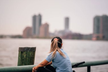 Genç bir kadın oturmuş deniz grisi gökyüzü ufku binalarını seyrediyor.